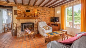 A cozy living room with exposed wooden beams, a brick fireplace, a television, and a large window with a view of greenery. - Charming cottage with enclosed garden in Green Picardy (Martincourt)