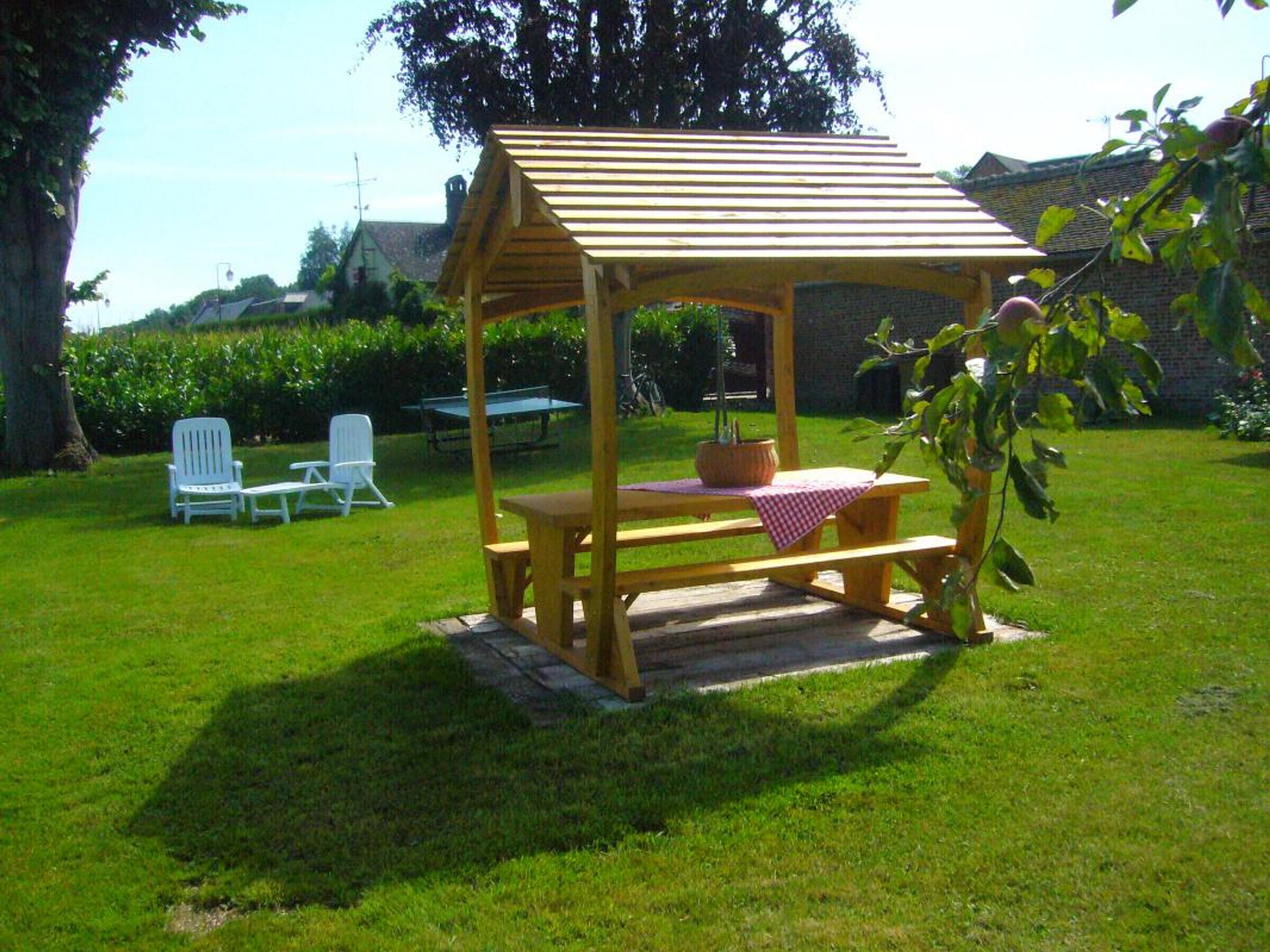 A wooden gazebo with a bench and picnic table, surrounded by a grassy lawn and trees.