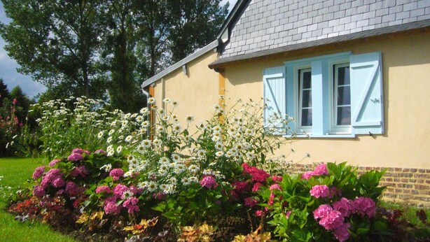 A house with a brick roof and light blue shutters, surrounded by a garden with various flowers and a large tree. - Charming cottage with enclosed garden in Green Picardy (Martincourt)
