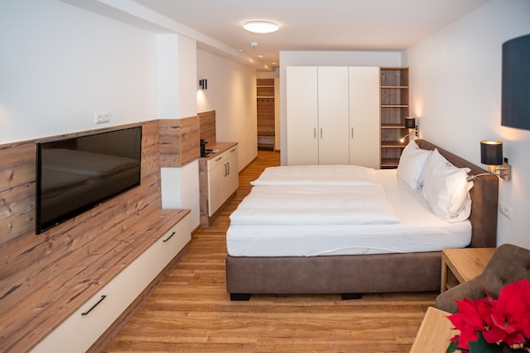 A modern bedroom with a large bed, a TV mounted on a wooden panel, a wardrobe, and a bedside table with a lamp.