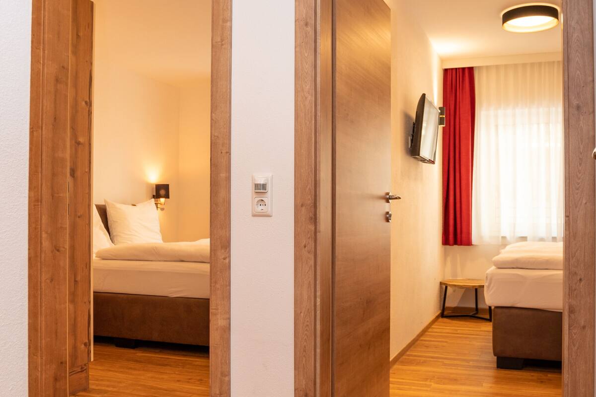 A hotel room with wooden flooring, a bed, a bedside table, a TV, and a window with curtains.