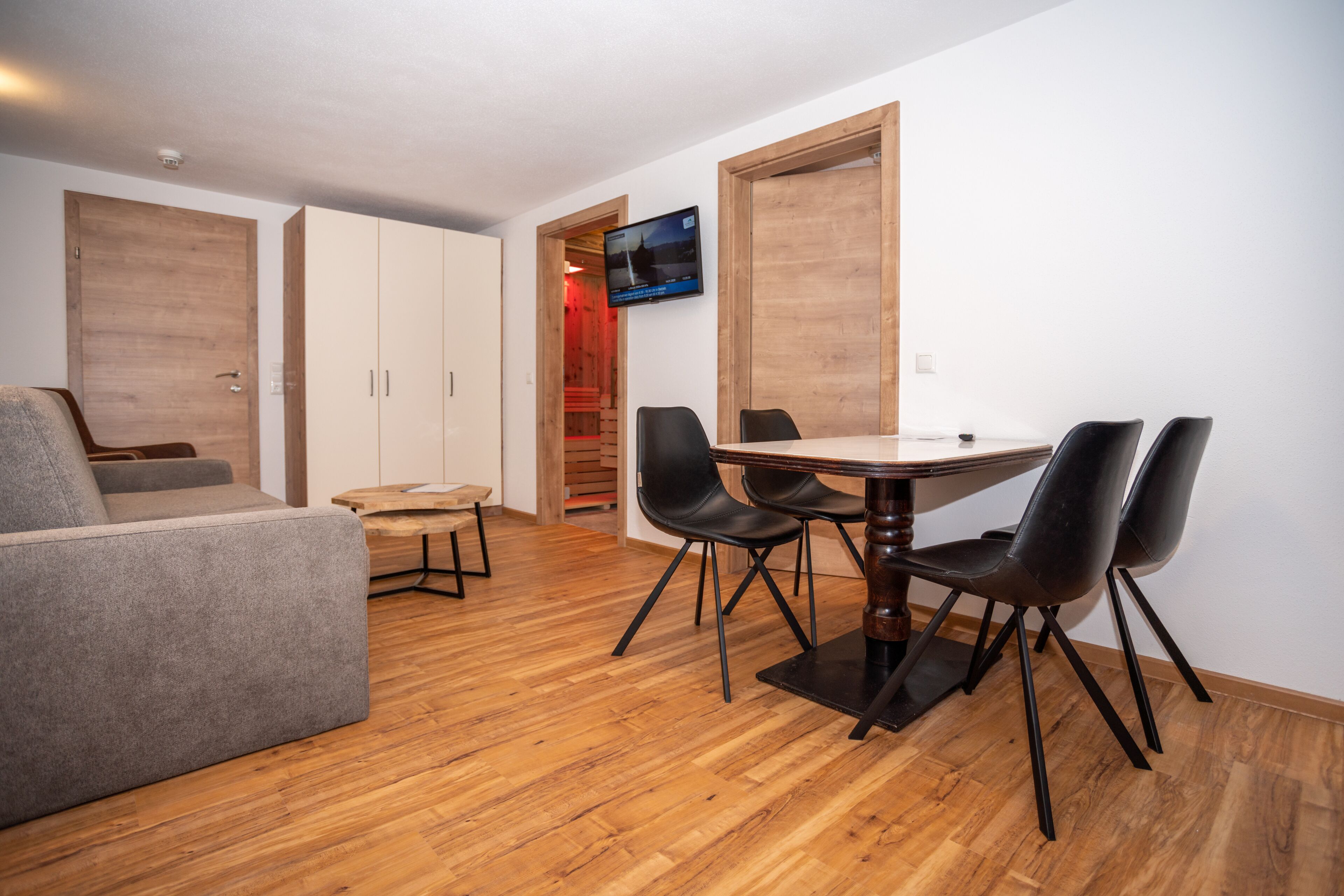 A modern living room with a sofa, a dining table with chairs, wooden flooring, and a TV mounted on the wall.