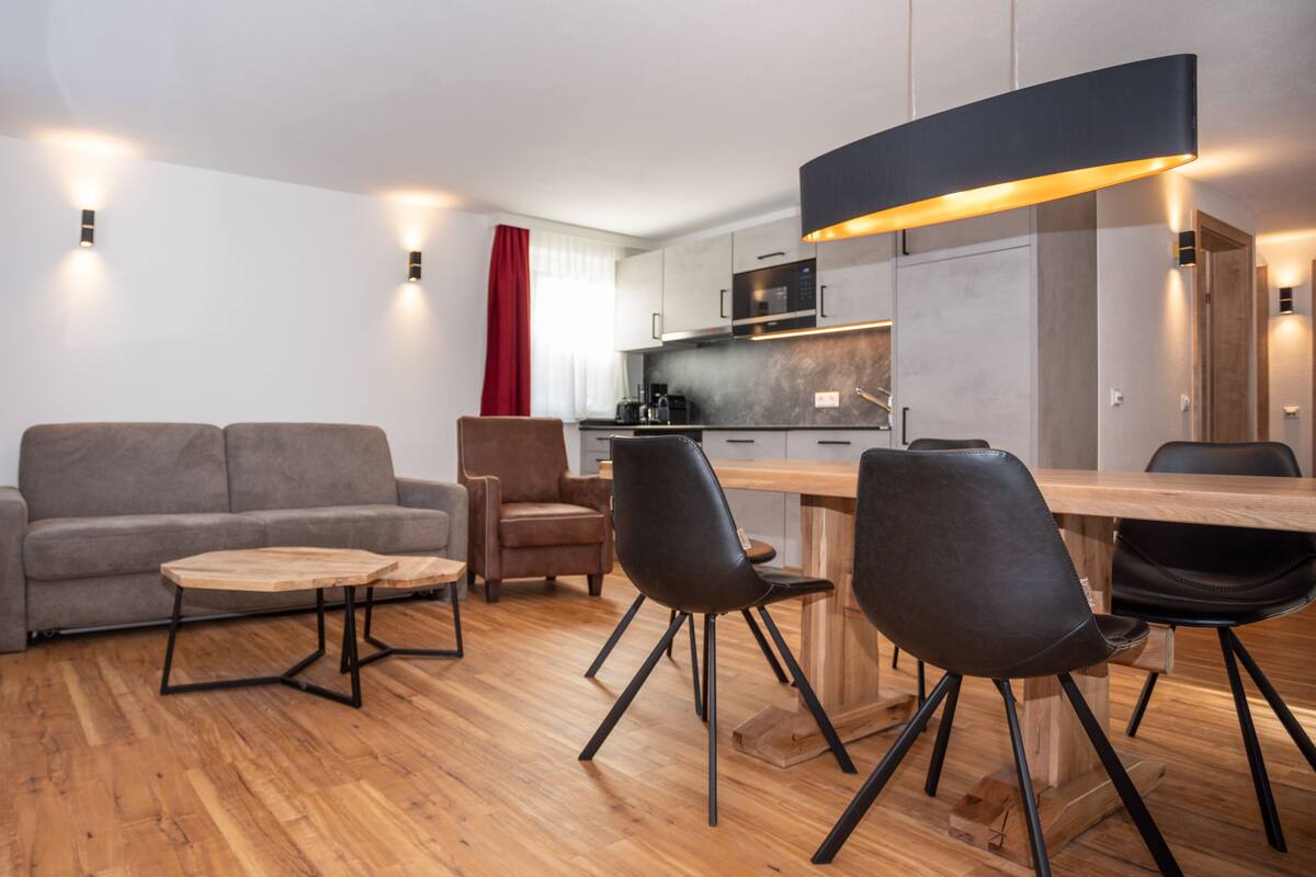 A modern living room with a sofa, two chairs, a wooden coffee table, and a dining table with chairs. There is a kitchen area with a microwave and cabinets.