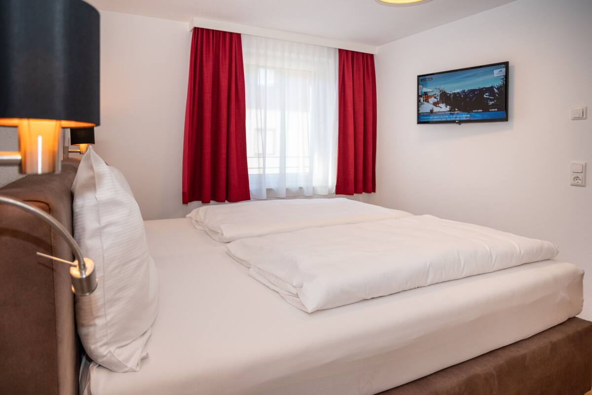 A hotel room with a bed, red curtains, a television on the wall, and a lamp on the left.