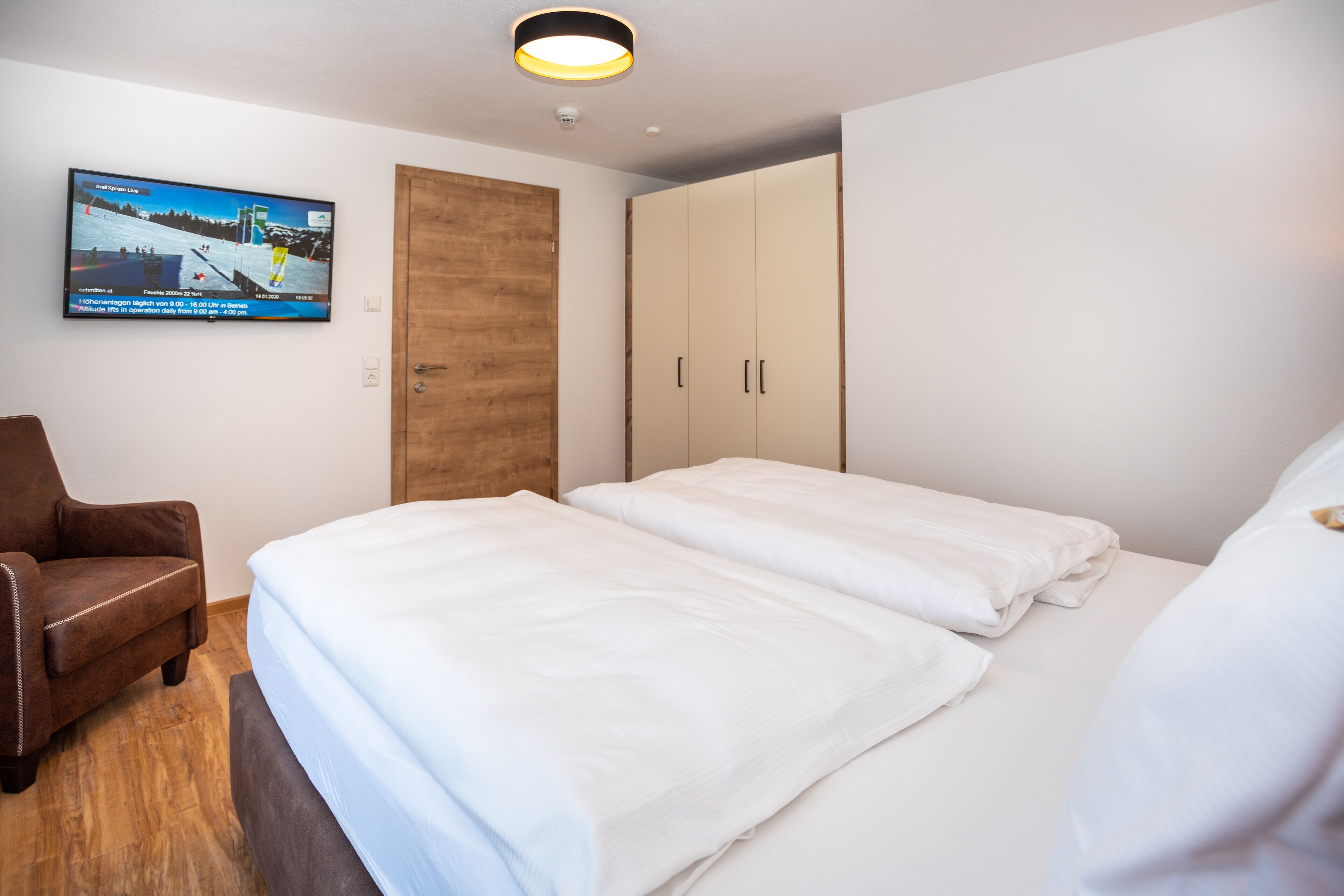 Une chambre d’hôtel avec un lit, une armoire en bois, un fauteuil marron et un téléviseur diffusant un événement sportif.