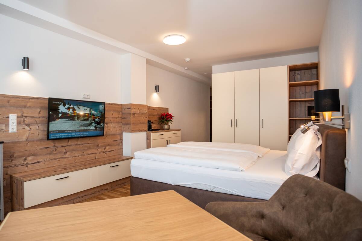 A modern bedroom with a wooden accent wall, a flat-screen TV, a bed with white linens, a bedside table, and a wardrobe.