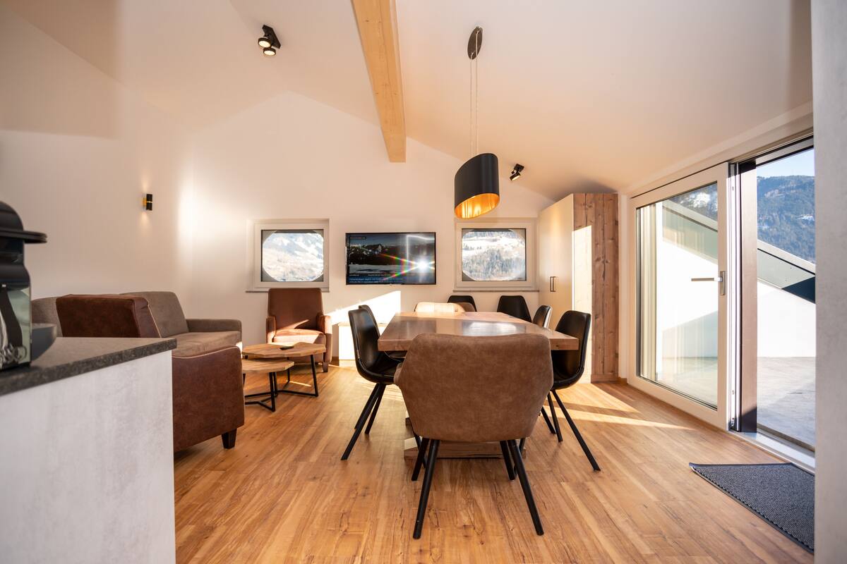 A modern dining area with a wooden floor, a round table, and several chairs. There are two wall-mounted televisions displaying scenic images.