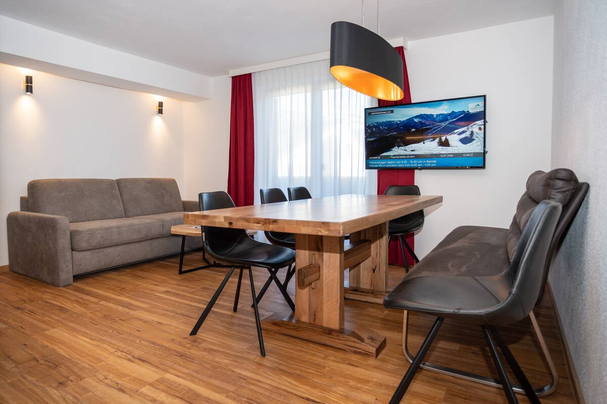 A modern living room with a wooden dining table, black chairs, a grey sofa, and a TV mounted on the wall displaying a snowy landscape.