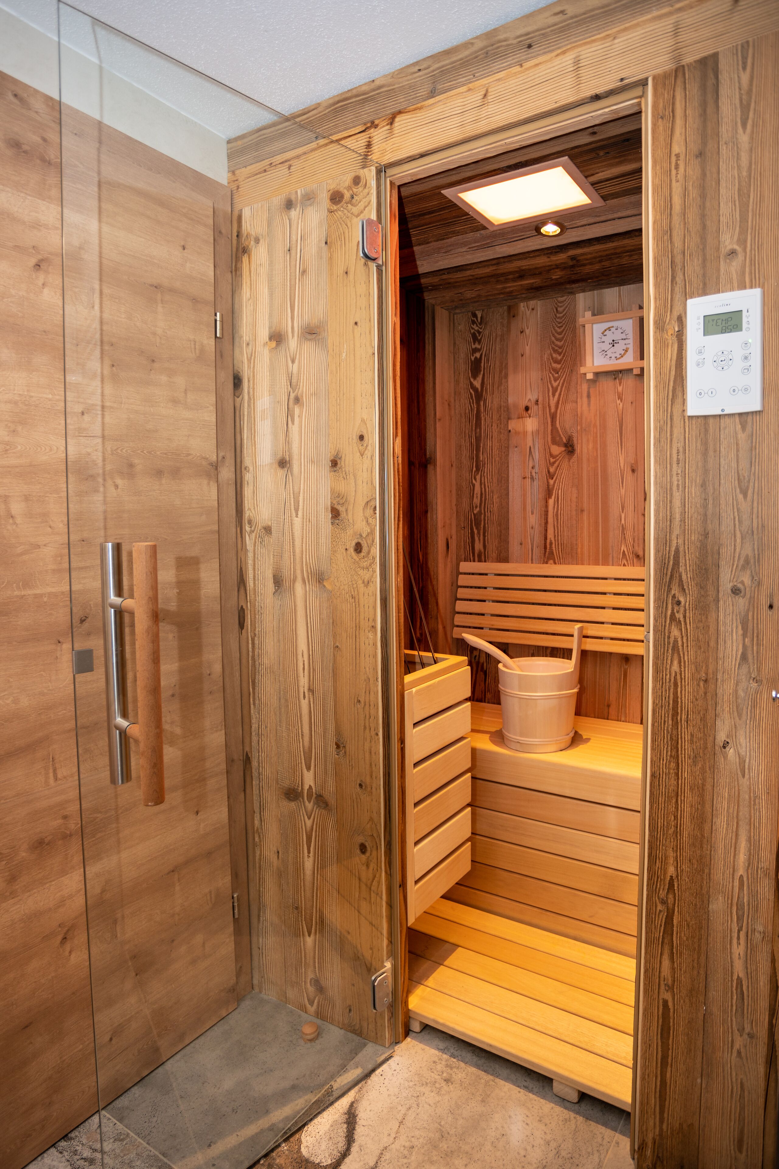 A wooden sauna with a bench, a bucket, and a control panel.