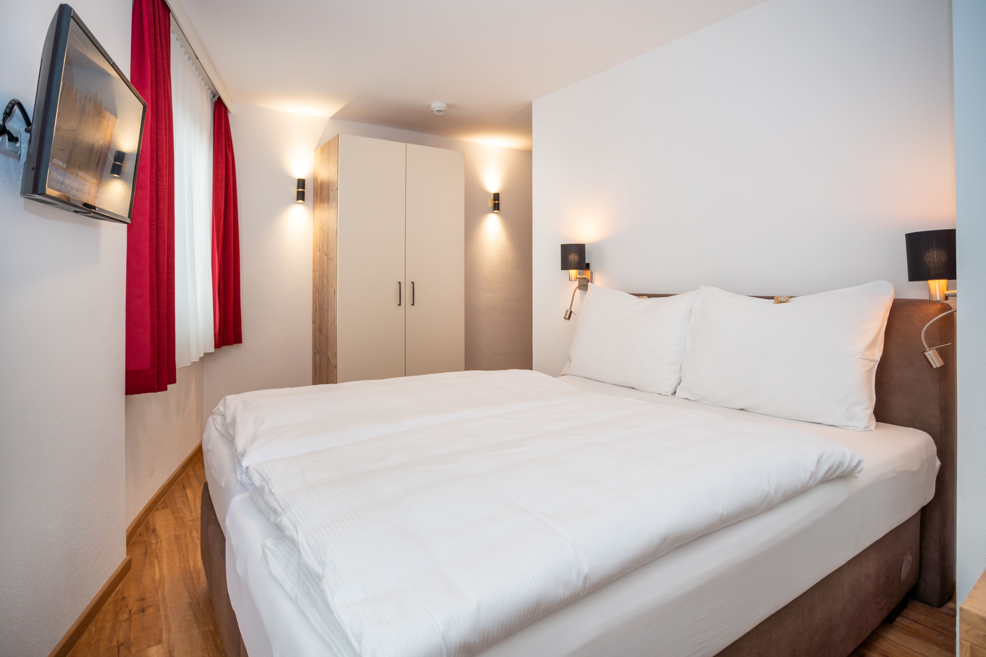 A neatly made bed in a hotel room with a wooden floor, a wardrobe, and two wall-mounted lamps.