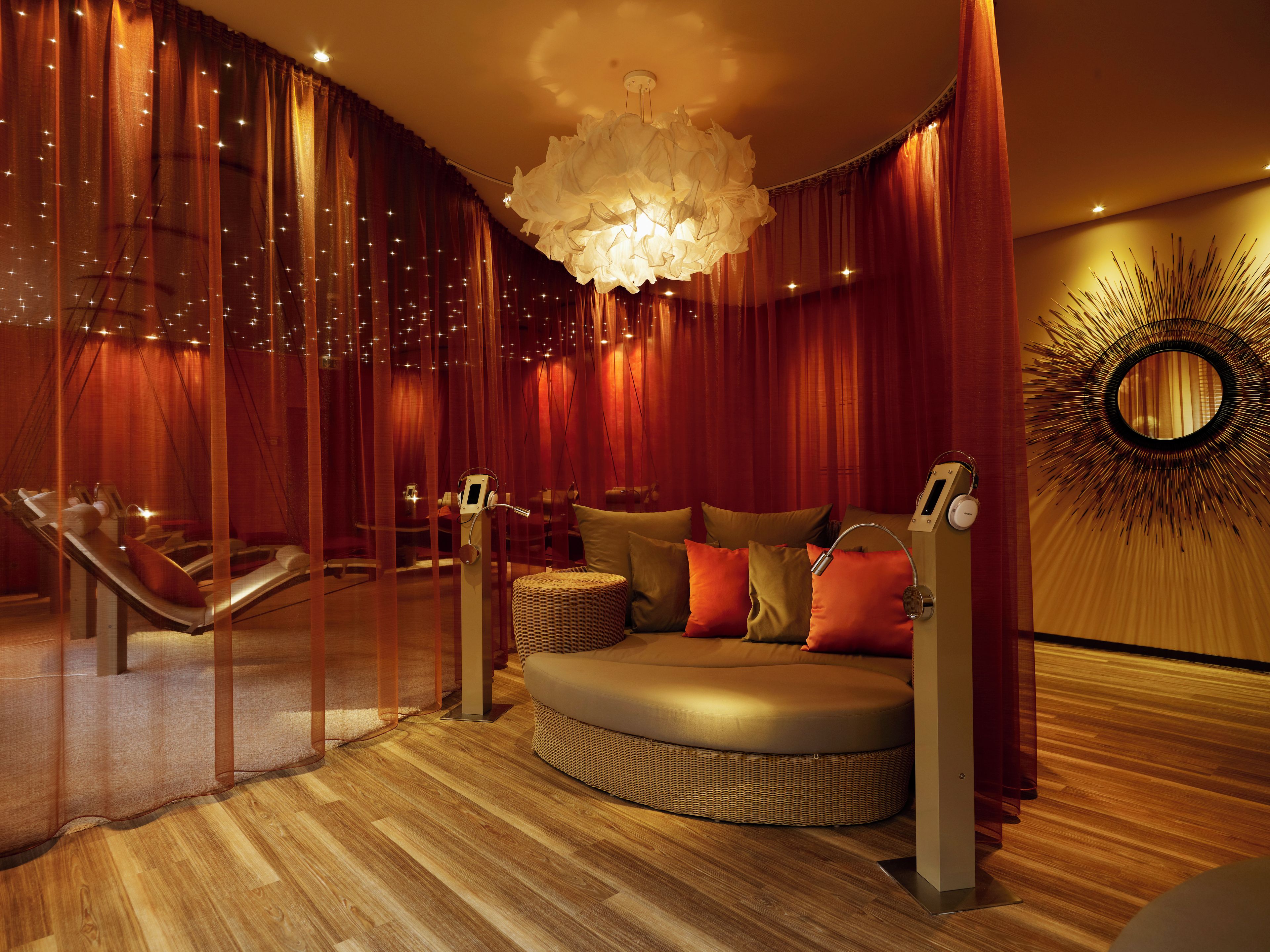 A room with a curved sofa, a massage chair, a large chandelier, and a wall with a sunburst design.