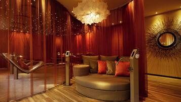 A room with a curved sofa, a massage chair, a large chandelier, and a wall with a sunburst design.