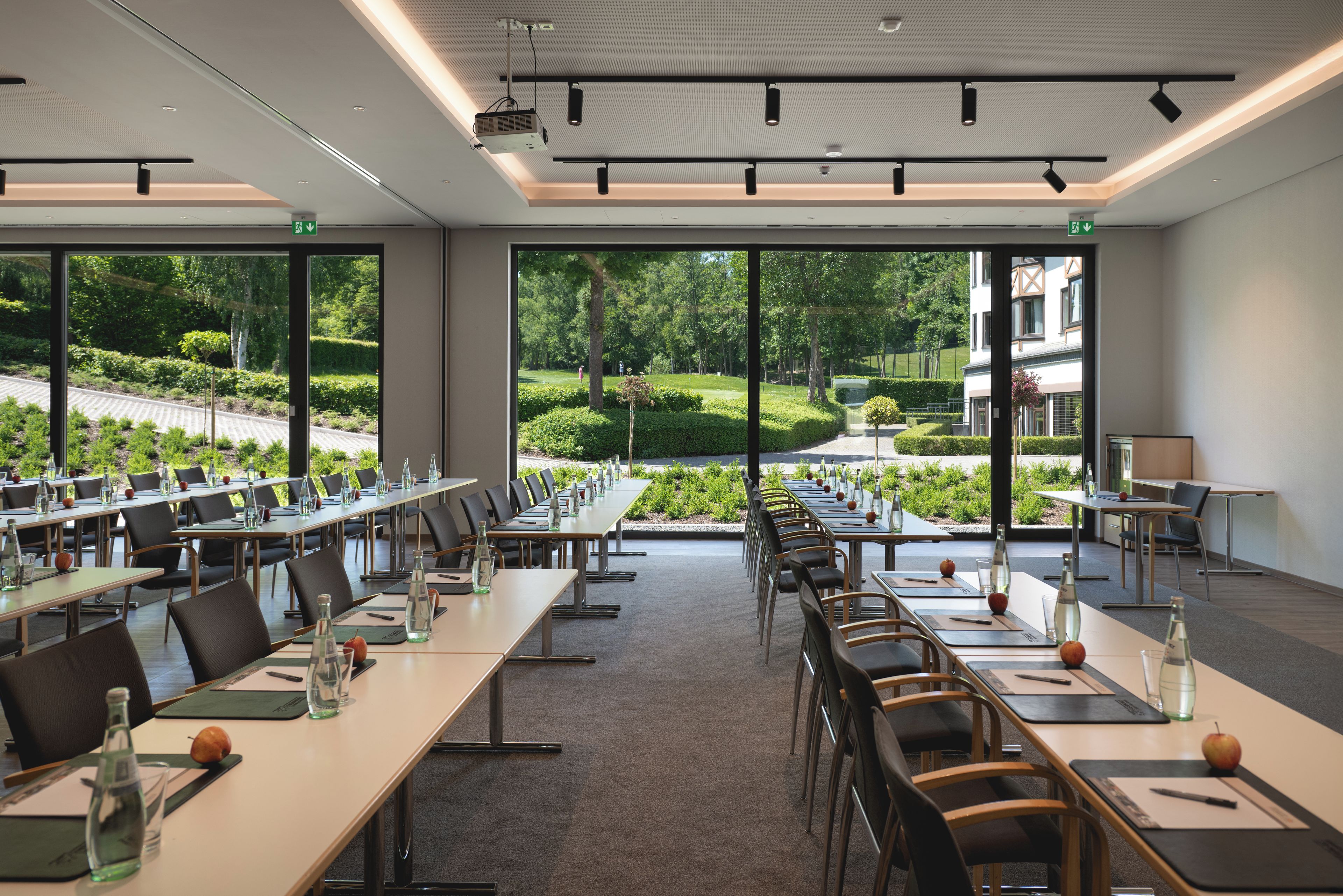 A conference room with long tables, chairs, and a view of a landscaped garden through large windows.