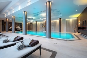 A modern indoor swimming pool with a clear water surface, surrounded by large pillars, and a bench with towels. - Land  & Golf Hotel Stromberg (Stromberg)