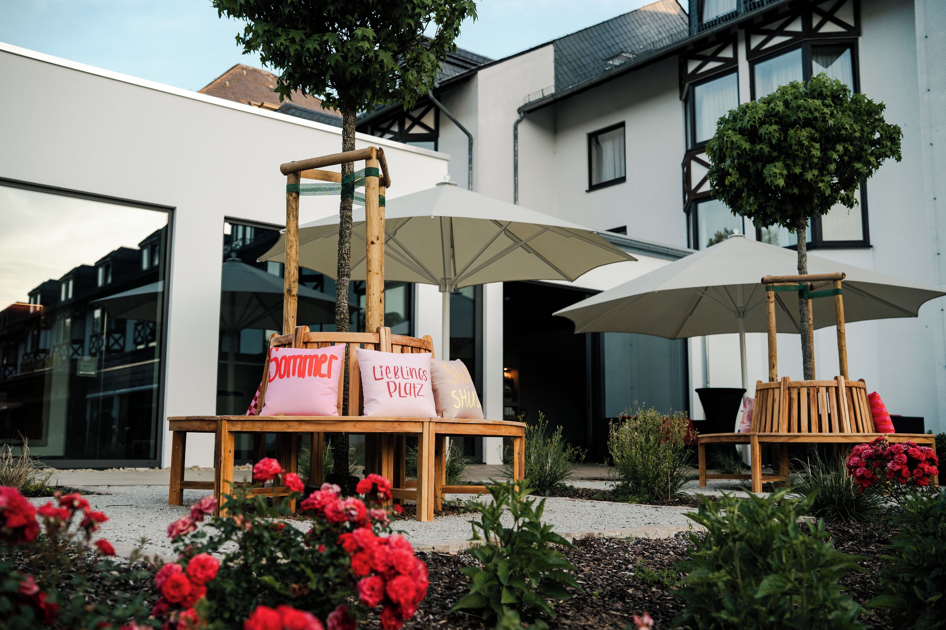 Terrasse mit Sonnenschirmen und Kissen, ein Baum und rote Blüten.