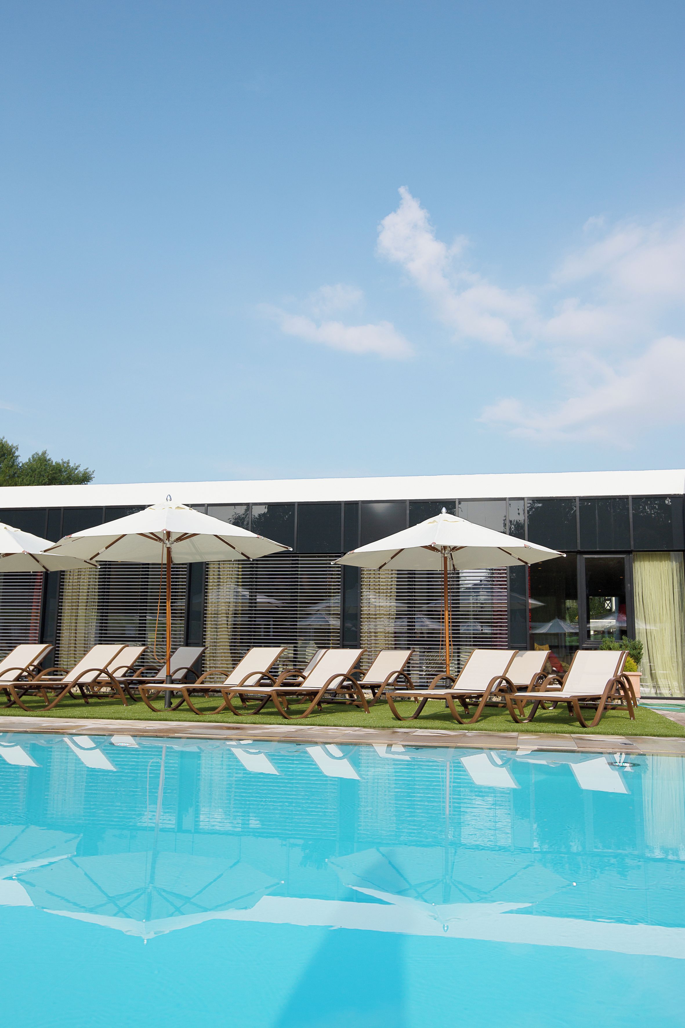Ein Schwimmbecken mit Liegestühlen und Sonnenschirmen, ein modernes Gebäude mit großen Fenstern und ein klarer Himmel.