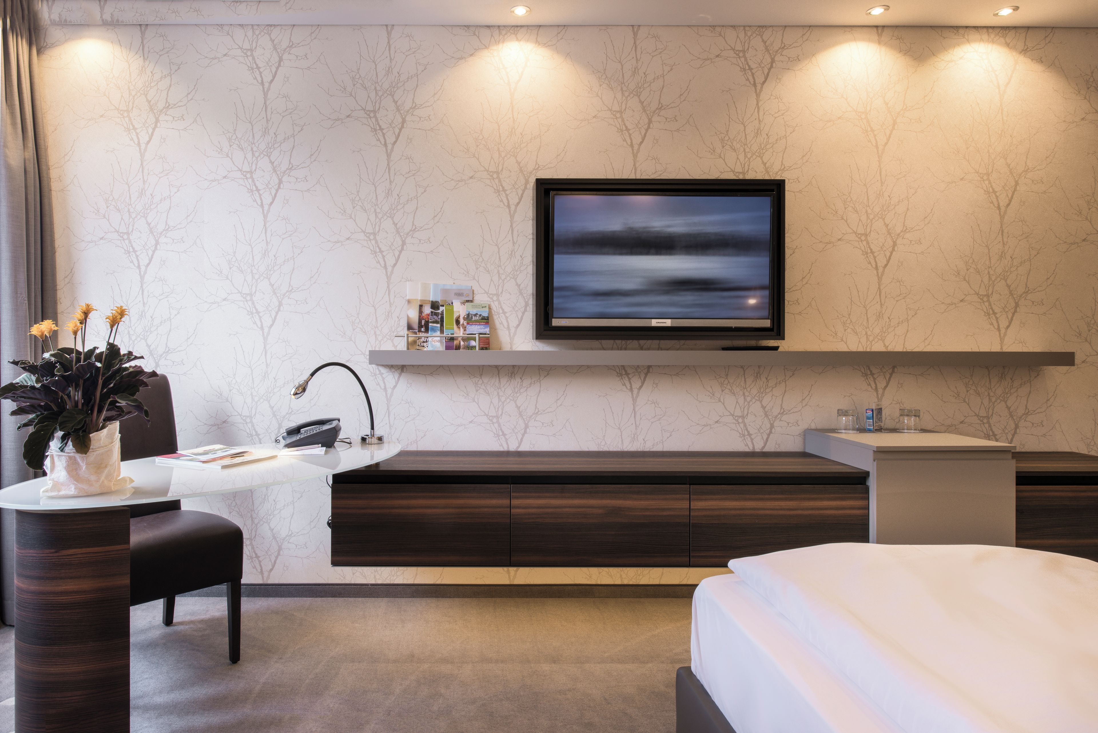 A modern hotel room with a large flat-screen TV mounted on the wall, a desk with a lamp, a chair, a bed, and a small side table with a vase of flowers.