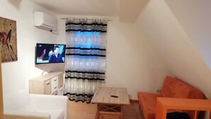A small, cozy living room with a white sofa, a wooden coffee table, and an orange armchair. A flat-screen TV is mounted on the wall, and a striped curtain covers the window. - In the house rose garden, center BT. 8 min. , Wifi - free parking, balcony, bus. (Bayreuth)