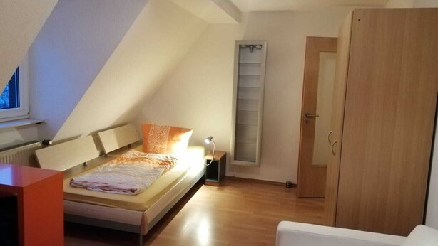 A bedroom with a sloped ceiling, a bed with a headboard, a bedside table, a nightstand, a window with blinds, and a wooden wardrobe. - In the house rose garden, center BT. 8 min. , Wifi - free parking, balcony, bus. (Bayreuth)