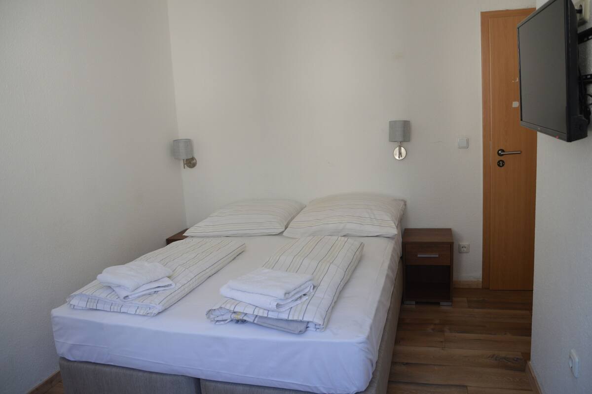 A neatly made bed with pillows and folded blankets, a bedside table, a wall-mounted TV, and a wooden door.