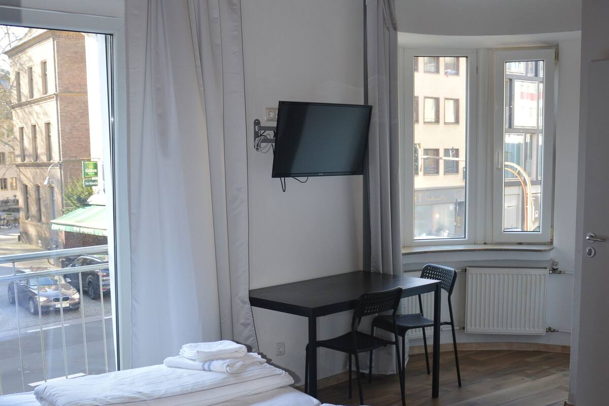 A hotel room with a bed, a desk, a chair, a TV, and a window with a view of the street.