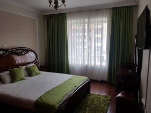 A hotel room with a large bed, green curtains, a wooden headboard, and a television on the wall. - Ken S Haven Riara Road (Nairobi)