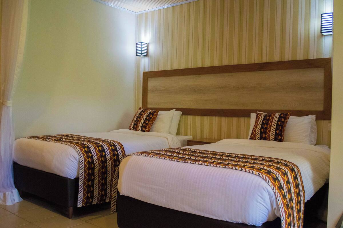 A hotel room with two beds, each with a patterned bedspread and pillows, a wooden headboard, and a wall-mounted light fixture.