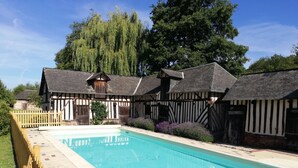 A swimming pool with a wooden deck and a building with a Tudor-style facade in the background. - Le Cottage de Cour la Ville (Le Mesnil-sur-Blangy)
