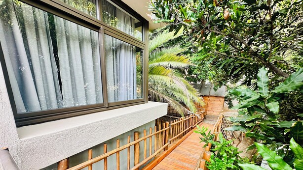 A wooden deck with a railing, a window with curtains, and lush greenery. - Little Forest Garden Retreat Guesthouse (Windhoek)