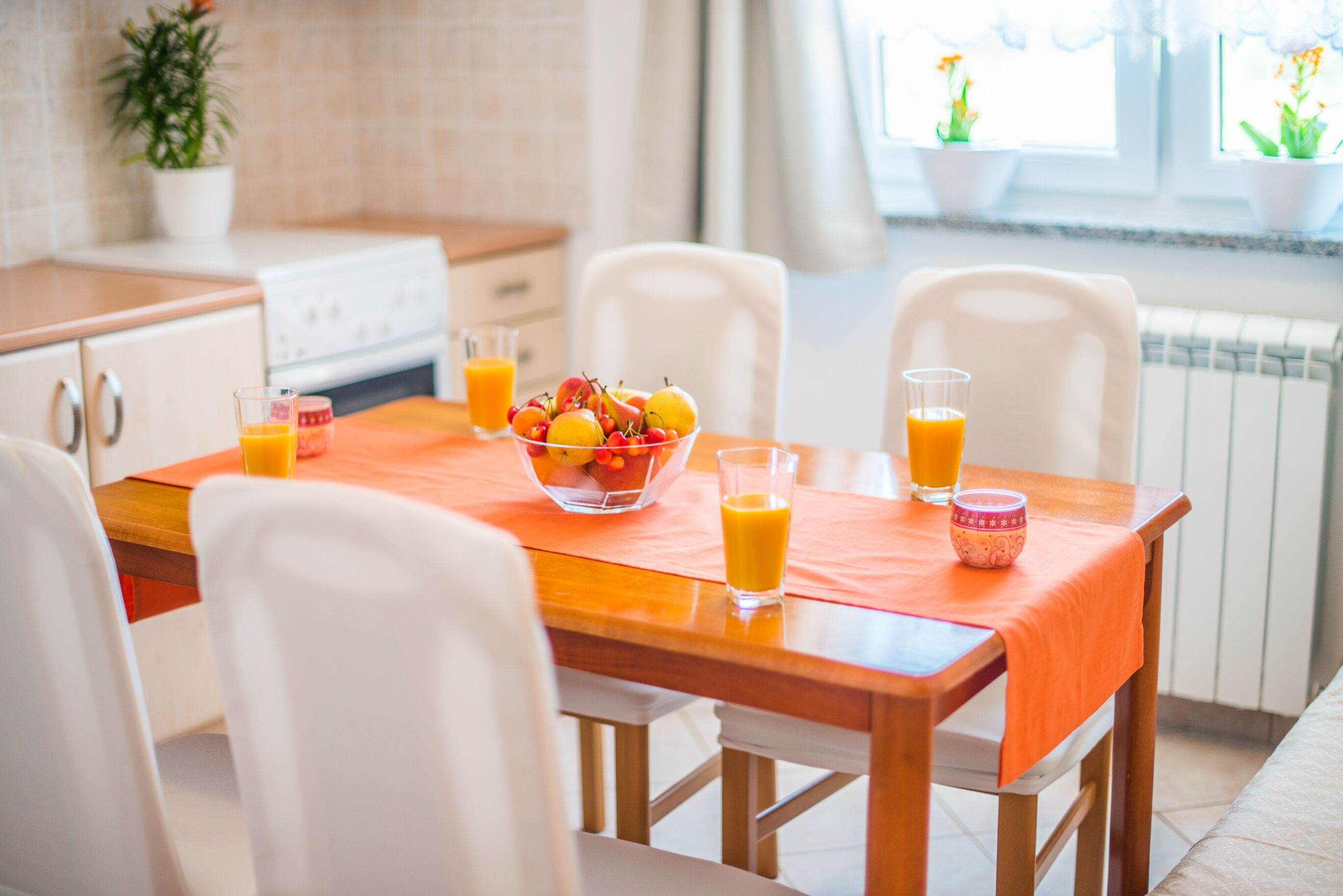 Une cuisine avec une table en bois, des chaises blanches, un saladier rempli de fruits et des verres de jus d’orange.