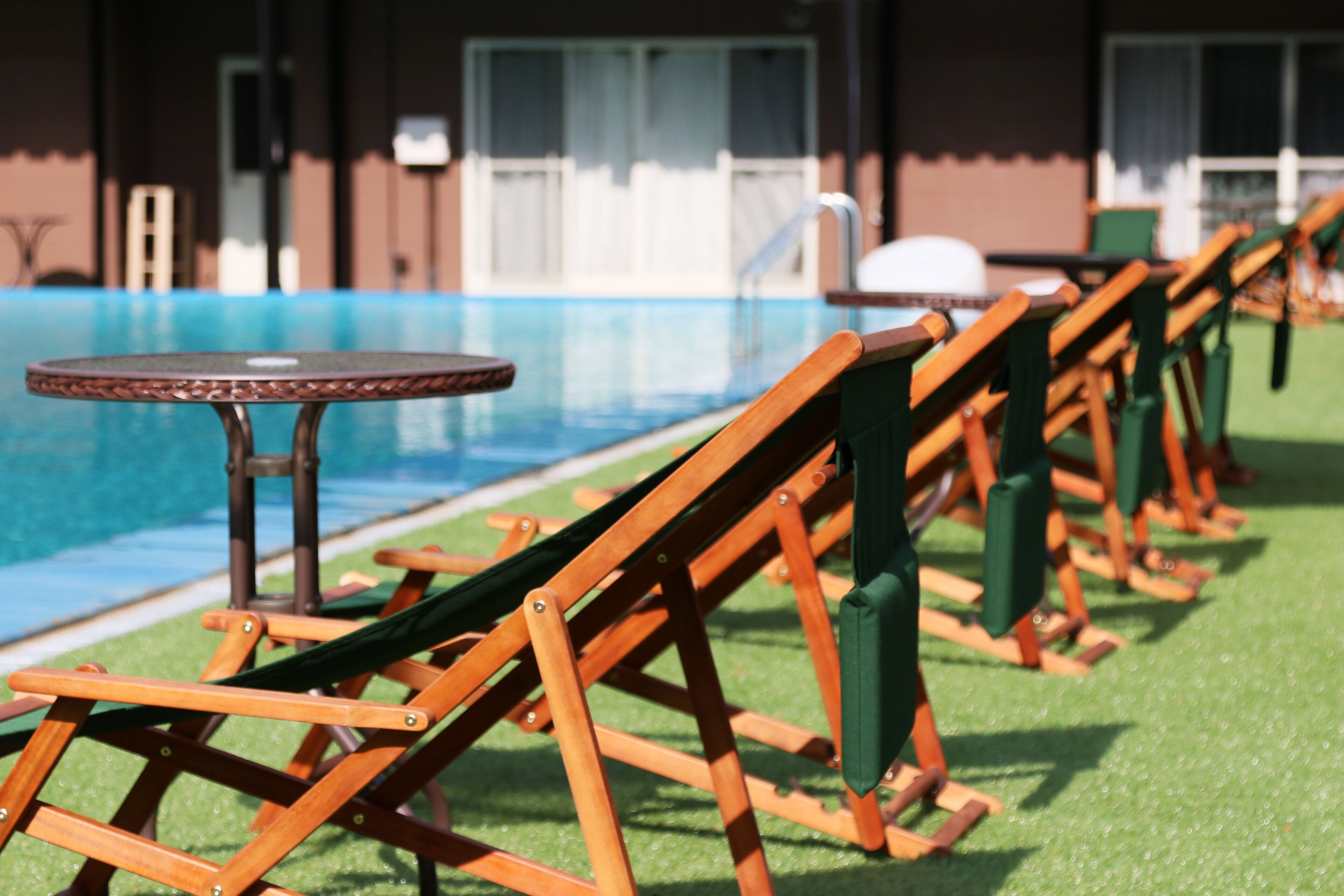 Filas de sillones y mesas de madera junto a una piscina, con un edificio al fondo.