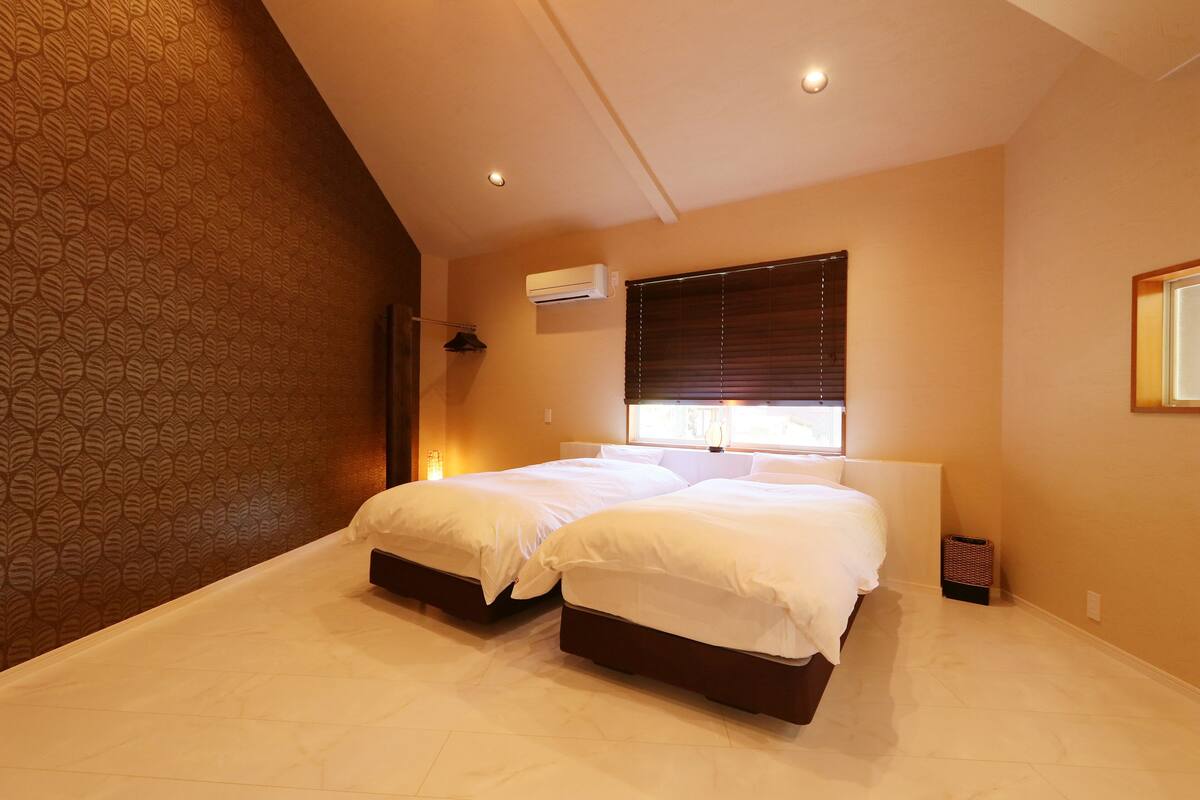 A bedroom with two beds, a window with blinds, an air conditioning unit, and a patterned wall.