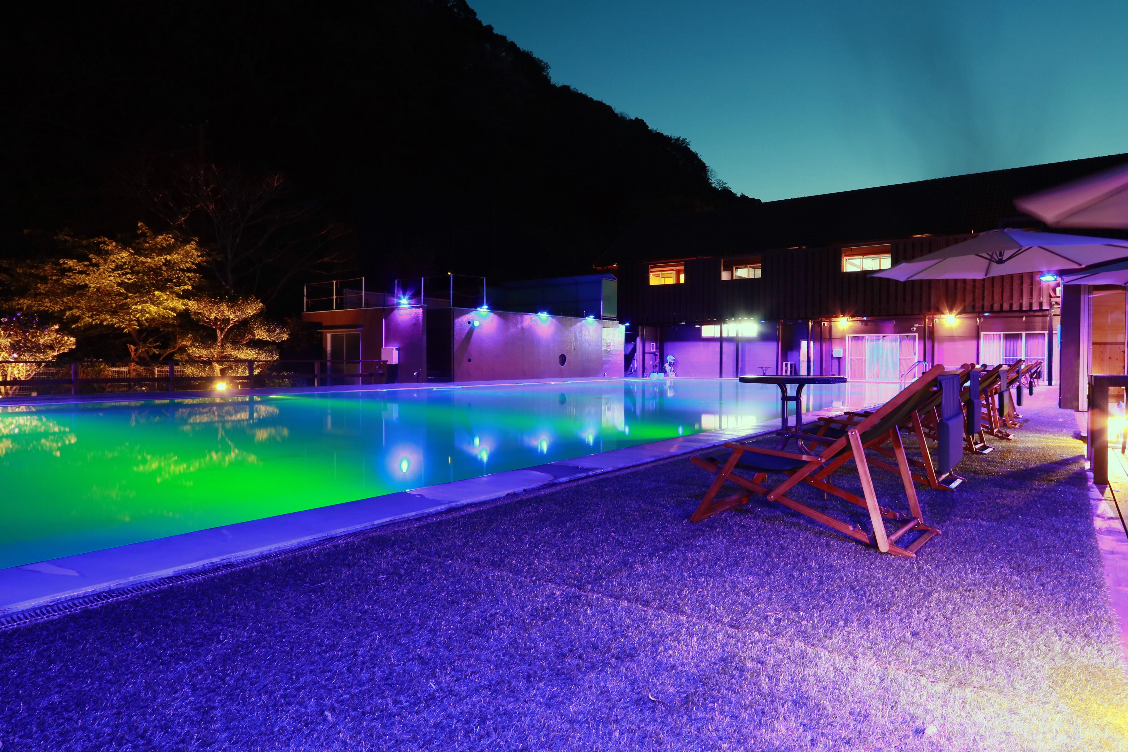 A swimming pool with a wooden deck and lounge chairs, surrounded by buildings with outdoor lighting.