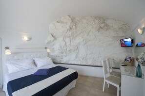 Un dormitorio con cama, cabecera blanca, pared con superficie texturizada en blanco, televisor, silla y escritorio con un jarrón.