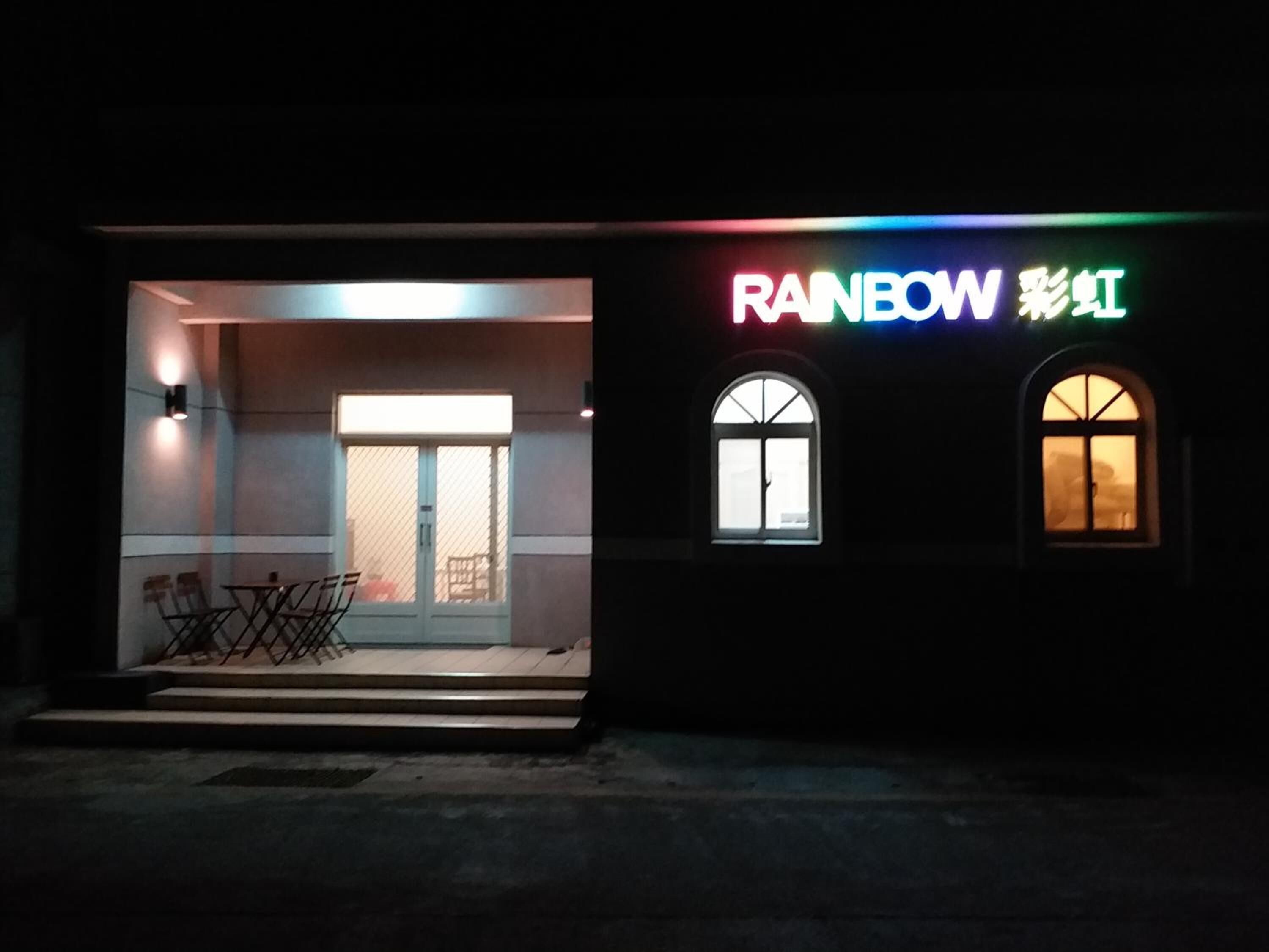 Um edifício com um letreiro de neon que diz 'RAINBOW' e caracteres chineses. Possui duas janelas e uma porta com painel de vidro.