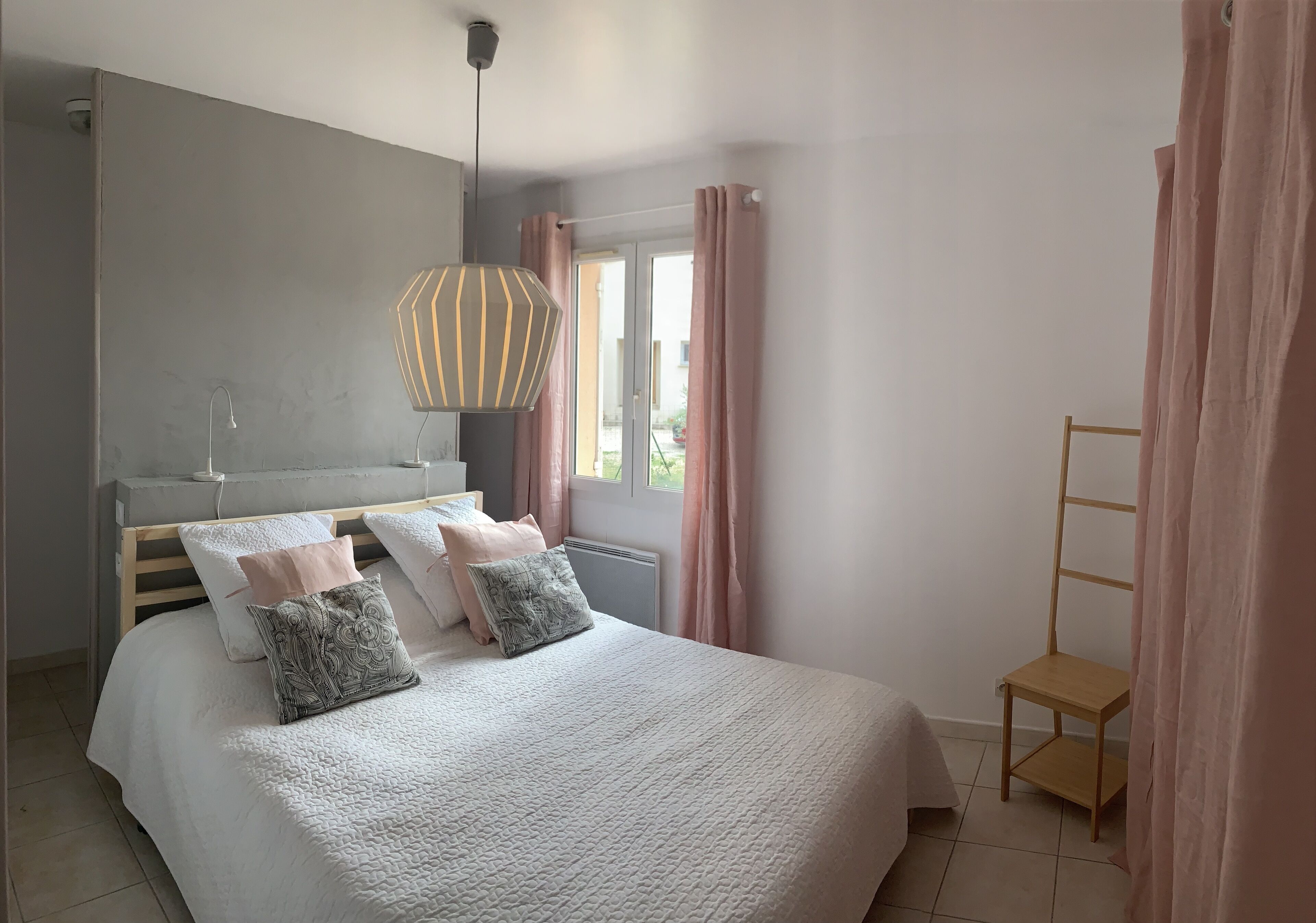 Une chambre à coucher comprenant un lit, une table de chevet, une lampe, une fenêtre avec des rideaux et une vue sur l’extérieur.
