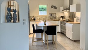 Cucina moderna con mobili bianchi, zoccolo in piastrelle e una finestra. Due sedie nere ad una tavola di legno, con una bottiglia di vino e un bicchiere.