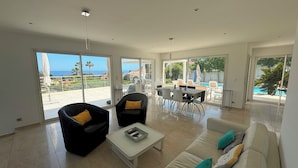 Ein großzügiges Wohnzimmer mit einer Couch, zwei Sesseln, einem Couchtisch und einem Essbereich mit Stühlen und einem Tisch. Große Schiebeglasfenster führen auf einen Balkon mit Blick auf das Meer und Palmen.