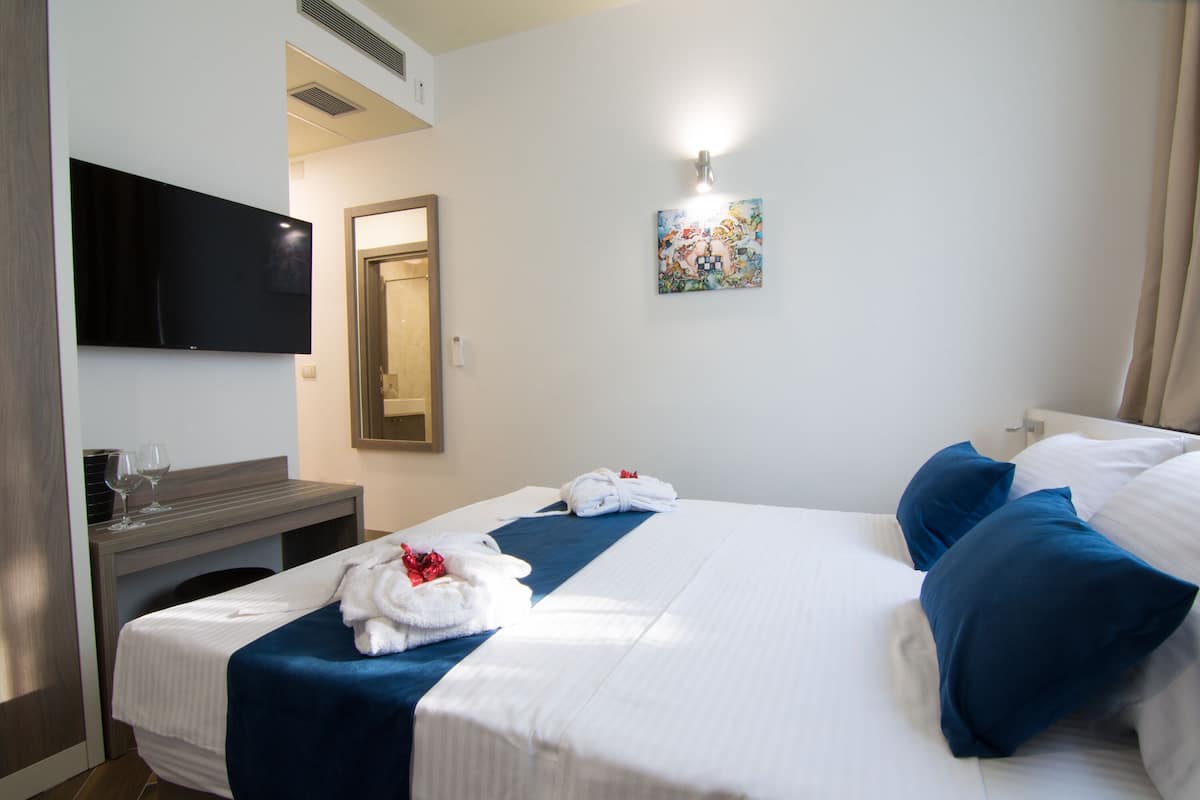 A hotel room with a bed, a desk with a flat-screen TV, a painting on the wall, and a window with curtains.
