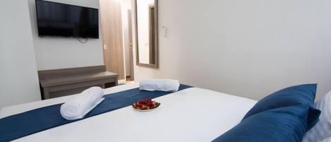 A hotel room with a bed, a TV mounted on the wall, a mirror, and a small table with a bowl of fruit.