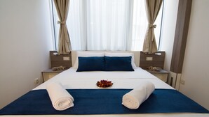 Cama arrumada com roupa de cama azul, uma tigela de frutas e toalhas dobradas em um quarto de hotel.