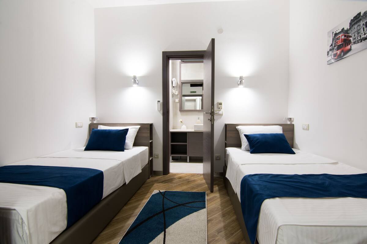 A hotel room with two single beds, a wooden floor, a small rug, and a door leading to a bathroom.