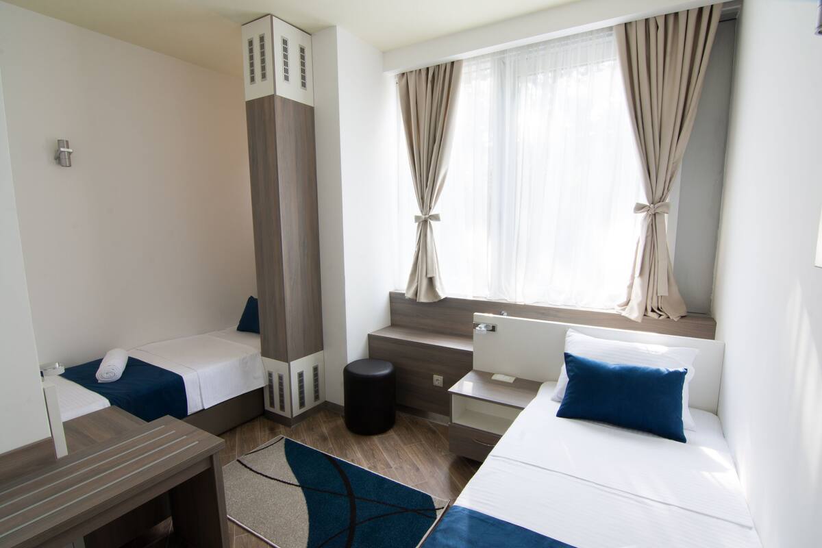 A hotel room with two beds, a desk, a small stool, and a window with curtains.
