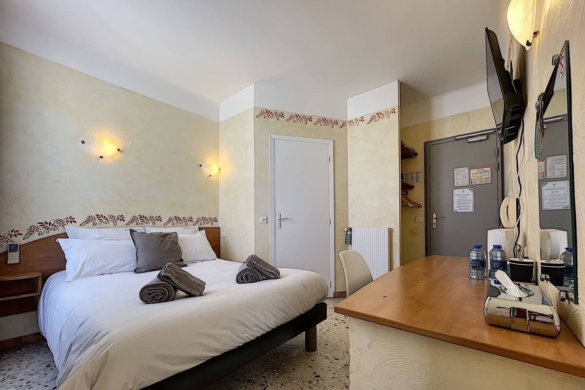 A hotel room with a bed, bedside table, and a desk with a chair. There are towels on the bed, a radiator, and a door with a mirror and papers on the wall.