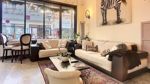 Un salon comprenant un canapé, des fauteuils, une table basse et une grande fenêtre donnant sur la rue.