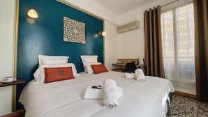 Une chambre d’hôtel avec un grand lit, deux oreillers, une table de chevet, une lampe, un élément décoratif mural, un climatiseur, un bureau, une chaise et une fenêtre avec des rideaux.