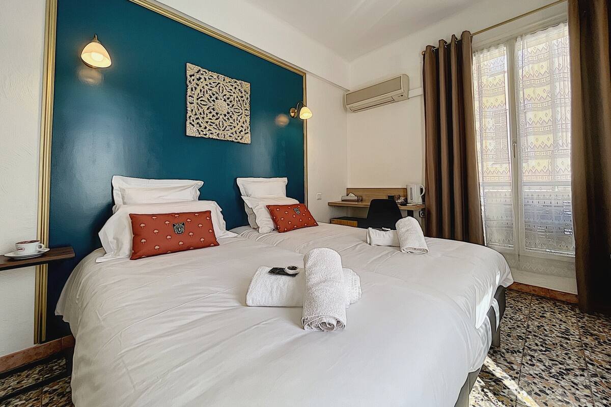 A hotel room with a large bed, two pillows, a bedside table, a lamp, a decorative wall piece, an air conditioning unit, a desk, a chair, and a window with curtains.