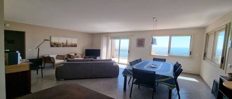 A spacious living area with a dining table, sofas, and a balcony with a view of the sea.