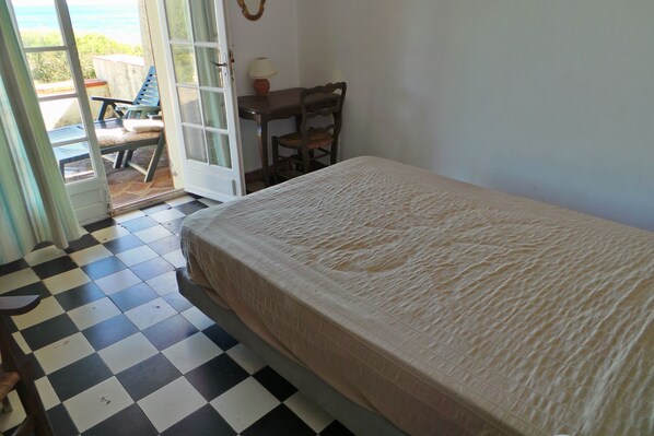 A bedroom with a checkered floor, a bed with a beige cover, a wooden table with a lamp, and a chair.