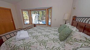 Une chambre avec un grand lit, une fenêtre donnant sur une maison et des arbres, une porte en bois et une table de chevet avec une lampe.